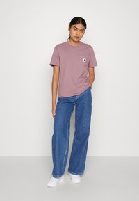 T-shirt color malva chiaro con taschino e logo. Abbinata a jeans blu a vita alta e sneaker bianche. Design semplice, vestibilità casual.