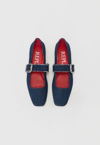 Chaussures en denim à bout carré avec une large sangle dotée d'une boucle en métal, semelles intérieures rouges et accents effilochés le long des bords de la sangle.