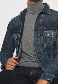 Jeansjacka i mörkblått med två bröstfickor, lätta slitage detaljer och silverfärgade detaljer, bärs över en grå polotröja.