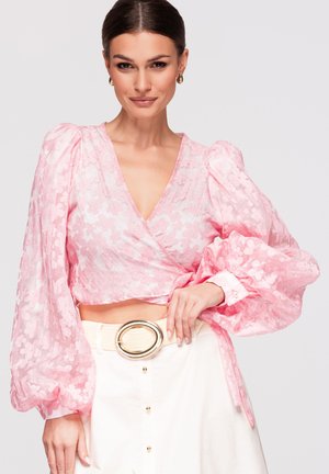 Femme aux cheveux foncés portant un chemisier portefeuille rose à fleurs avec des manches bouffantes et une jupe blanche avec une large ceinture à boucle dorée, souriant légèrement.