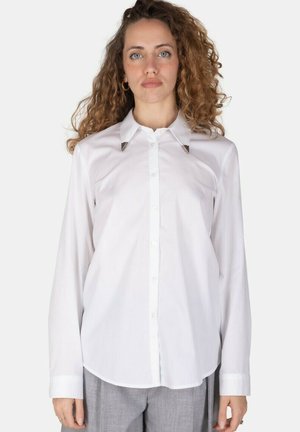 Donna con capelli ricci indossa una camicia bianca a maniche lunghe con punte del colletto argentate e pantaloni grigi, in piedi davanti a uno sfondo semplice.