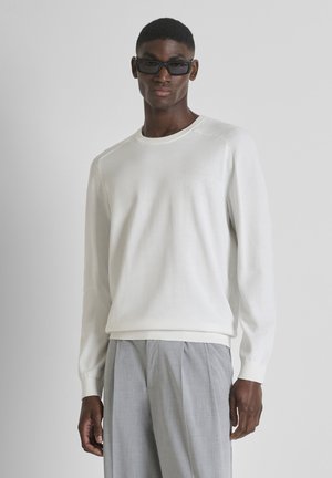 Man met zwarte zonnebril, witte trui met lange mouwen en lichtgrijze geplooide broek staand tegen een effen grijze achtergrond.