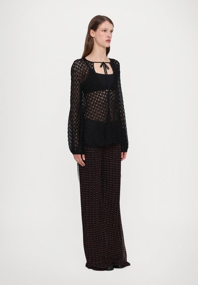 Blusa nera trasparente con un motivo testurizzato, dotata di un nodo frontale e maniche lunghe, indossata con pantaloni larghi a righe neri e rossi.