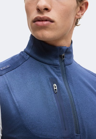 Gros plan d'une personne portant une veste bleue à col montant avec deux fermetures éclair visibles à l'avant et un logo RLX sur l'épaule.