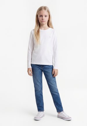 Lyshåret pige står iført hvid langærmet skjorte, blå jeans og hvide sneakers på en ensfarvet hvid baggrund.