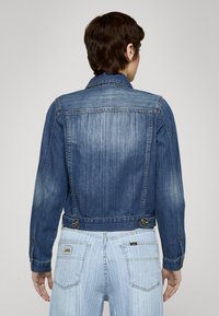 Rifľová bunda, stredne modrá, priliehavý strih, dva gombíky na páse, viditeľné švy, klasický golier a predný diel so záševkami.