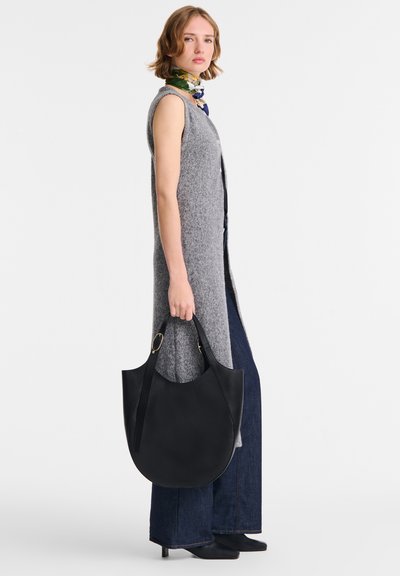 Frau trägt einen ärmellosen grauen langen Weste, dunkle weitgeschnittene Jeans, ein gemustertes Halstuch und hält eine große schwarze Ledertasche.