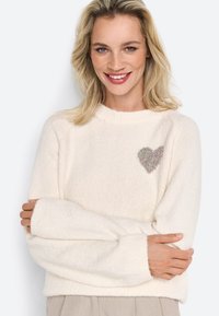 Bílý pletený svetr s volným střihem, který má na levé hrudi texturovaný design srdce a žebrované manžety. Měkký a útulný materiál.