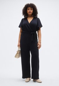 Fekete, wrap stílusú overál rövid, fodros ujjakkal, megkötős derékövvel és bő szárakkal. Világos bézs kézitáskával és krémszínű cipővel párosítva.