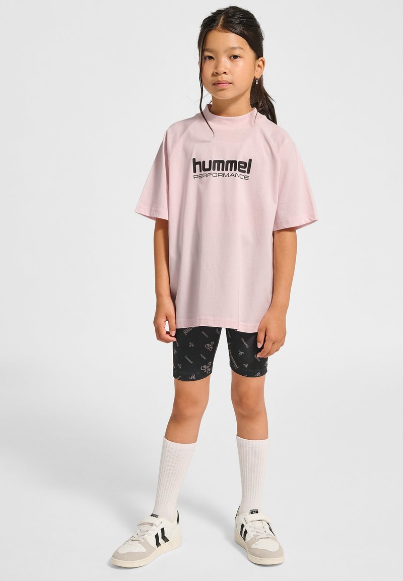 Różowa, oversize'owa koszulka z czarnym logo "hummel PERFORMANCE", połączona z czarnymi szortami z powtarzającym się wzorem graficznym, białymi skarpetkami i sportowymi butami.