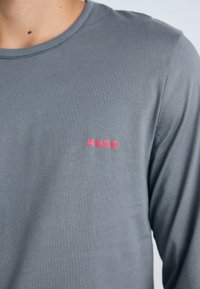 T-shirt gris à manches longues en coton avec un col rond. Présente un logo rouge "HUGO" positionné sur la poitrine à gauche.