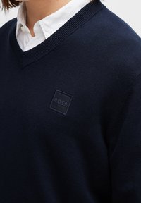 Tmavě modrý svetr s výstřihem do V, vyrobený z pletené látky. Na levé straně hrudi má texturovanou čtvercovou nášivku s nápisem "BOSS".