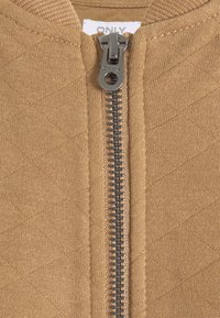 Tissu de veste matelassée brun avec une fermeture éclair argentée et des détails gris. Présente un col côtelé et une fermeture à glissière. Motif texturé sur l'ensemble.