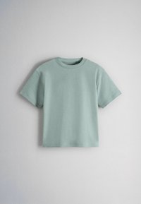 Világoszöld rövid ujjú pamut póló kerek nyakkivágással, egyenes felületre helyezve, sima világosszürke háttér előtt.