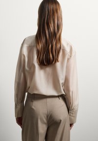 Blouse en soie beige clair avec une coupe décontractée, manches longues, poignets boutonnés, associée à un pantalon en toile beige. Texture lisse, détails minimaux.