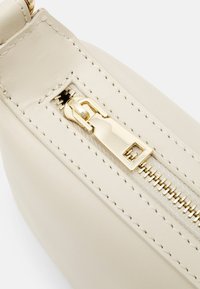Borsa in pelle beige con zip dorata e un tirante rettangolare, con cuciture visibili lungo i bordi. Dettaglio della superficie testurizzata.