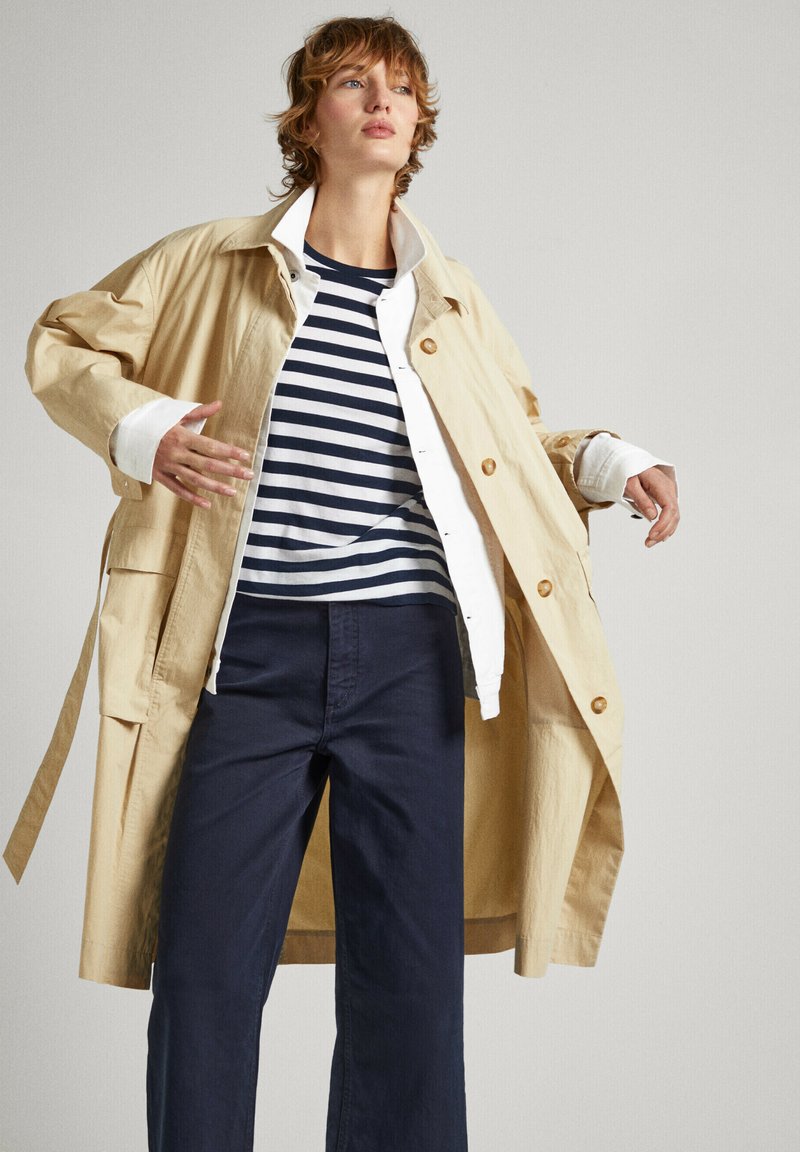 Manteau beige à grands boutons, porté sur une chemise blanche et un top à rayures marines, associé à un pantalon large marine. Texture lisse, coupe décontractée.