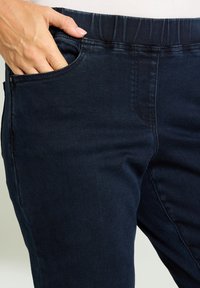 Mano con unghie curate poggiata all'interno della tasca anteriore di jeans elasticizzati blu scuro indossati con una maglietta bianca.