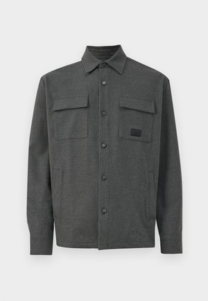 Grå skjorte med knapper laget av teksturert stoff, som har to frontlommer med klaffelukking og et lite logo-patch på brystet.