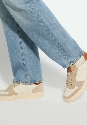 Jean bleu clair porté avec des baskets décontractées beiges et blanches, montré de la partie inférieure de la jambe jusqu'aux pieds sur un fond uni.