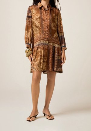 Femme portant une robe boutonnée à manches longues à motif cachemire marron, avec des bracelets dorés et des sandales à talons ouvertes marron.