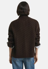 Persoon met kort, donker haar, die een donkerbruine kabelgebreide trui over een blauw-wit gestreept shirt draagt, gecombineerd met blauwe jeans, weggekeerderd.