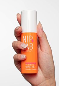 Bouteille cylindrique orange avec un bouchon à pompe blanche. L'étiquette comprend "NIP+FAB" et "SÉRUM VITAMINE C FIX 5%" en texte gras. Surface texturée, tenue dans une main.