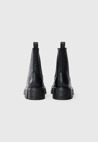 Botas de tornozelo em couro preto com sola de borracha grossa, inserts laterais elásticos e alças traseiras com texto em relevo no calcanhar.