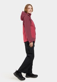 Veste à capuche bordeaux avec des manches contrastantes, poches zippées, portée sur un pantalon cargo noir et des baskets noires. Texture de tissu lisse.