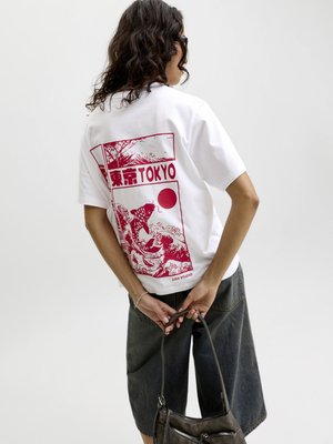 Personne aux cheveux bouclés portant un T-shirt blanc avec un motif rouge Tokyo et un short foncé, tenant un sac à main noir derrière le dos.