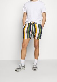 Raidalliset uimapuvut sinisessä, keltaisessa, vihreässä ja valkoisessa; joustava vyötärö, jossa naru; polvipituinen. Yhdistetty yksinkertaisen valkoisen t-paidan ja harmaiden lenkkareiden kanssa.