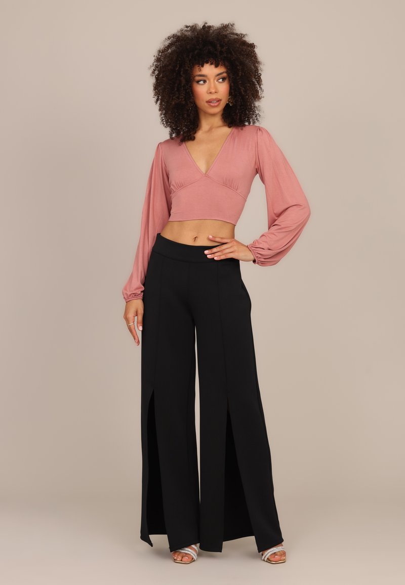 Γυναίκα με σγουρά μαλλιά που φοράει ένα ροζ crop top με μακριά μανίκια και μαύρο φαρδύ παντελόνι με ψηλή μέση και εμπρόσθιες σχισμές, στέκεται μπροστά σε ένα μπεζ φόντο.