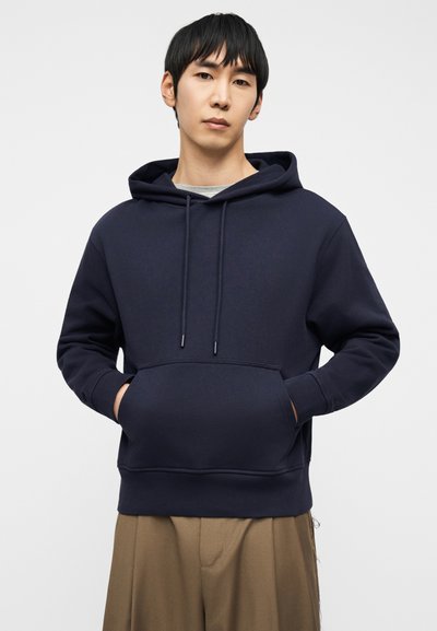 Νεαρός άνδρας που φοράει σκούρο μπλε φούτερ με κουκούλα, με τα χέρια στην μπροστινή τσέπη και μπεζ παντελόνι φαρδιάς γραμμής, μπροστά σε απλό φωτεινό φόντο.