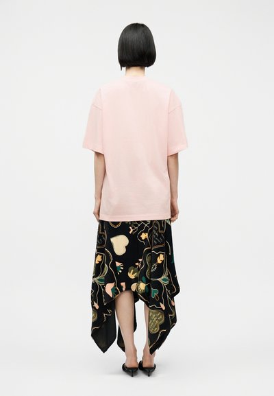 Άτομο με κοντά μαύρα μαλλιά που φοράει ένα ανοιχτό ροζ φαρδύ μπλουζάκι και μια μαύρη ασύμμετρη φούστα με μοτίβα καρδιών και λουλουδιών, γυρισμένο από πίσω.