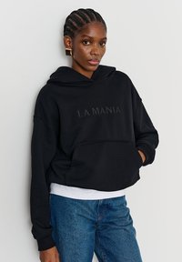 Hanorac negru scurt, cu buzunar frontal și text embosat "LA MANIA." Purtat peste o cămașă albă, asortat cu blugi albaștri din denim. Material neted.