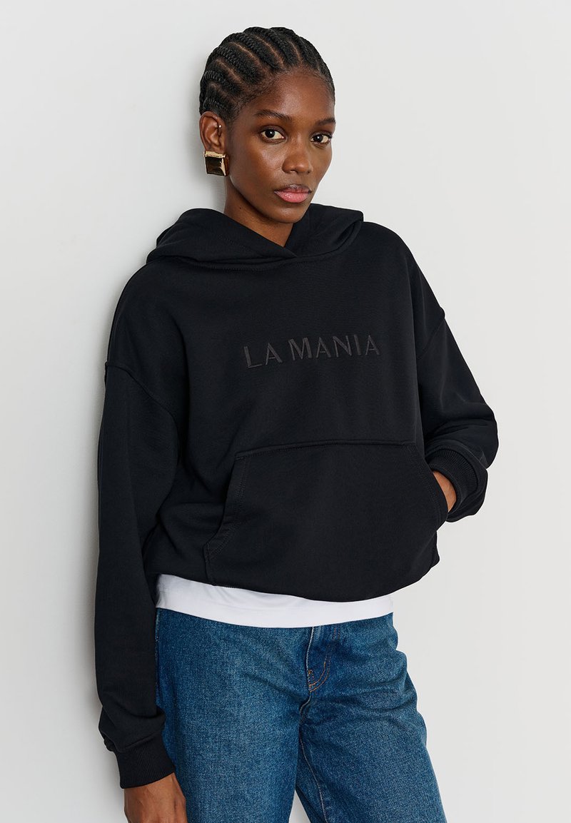 Hanorac negru scurt, cu buzunar frontal și text embosat "LA MANIA." Purtat peste o cămașă albă, asortat cu blugi albaștri din denim. Material neted.