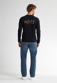 Zwart T-shirt met lange mouwen en gouden tekst en logo op de achterkant. Gekocht met lichtblauwe jeans en witte sneakers.