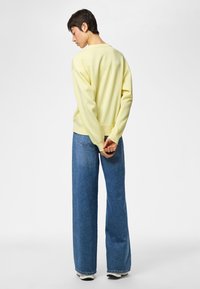 Persoon met kort donker haar, gekleed in een lichtgele trui, wijde blauwe spijkerbroek en witte schoenen, staand met de handen achter de rug in elkaar gestrengeld.