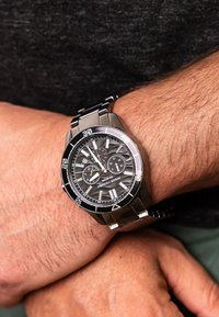 Nerūdijančio plieno laikrodis su tamsiai pilku tekstūruotu ciferblatu, sidabriniais valandų žymekliais, chronografų subdijais ir metaliniu apyrankiu.