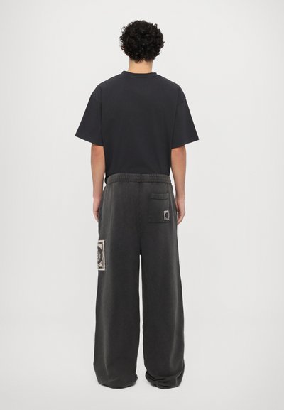 Grå brede bukser med struktureret overflade, elastisk talje og to baglommer med dekorative lapper. Sort t-shirt med korte ærmer.