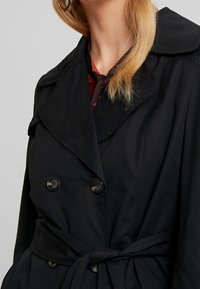 Czarny płaszcz trencz o kroju dwurzędowym, z dużym kołnierzem, paskiem do wiązania w talii oraz guzikami o lekko błyszczącej fakturze.