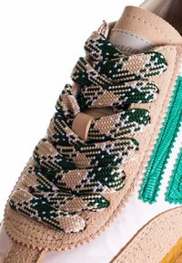 Sneaker con una toma superiore in suede beige, lacci a motivi verdi e bianchi, e una striscia verde testurizzata lungo il lato.