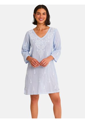 Femme souriante portant une robe bleu clair à col en V avec broderie florale blanche et rayures verticales, debout, mains jointes devant.
