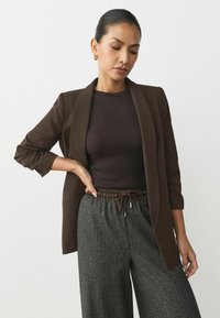 Sacou maro, ajustat, cu guler cârligat, purtat cu un top închis, cu fante ribbate, și pantaloni largi gri, texturați, cu talie mare.