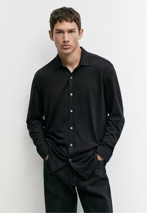 Giovane uomo con capelli corti che indossa una camicia nera abbottonata e pantaloni scuri, in piedi con le mani in tasca davanti a uno sfondo semplice.