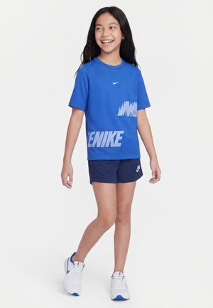 Smilende pige med langt mørkt hår iført en blå Nike T-shirt, navy shorts og hvide sneakers, stående foran en ensfarvet baggrund.