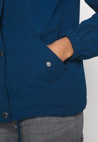 Osoba w niebieskiej kurtce z elastycznymi mankietami i szarymi spodniami, z ręką wsuniętą w przednią kieszeń zapinaną na metalowy guzik.