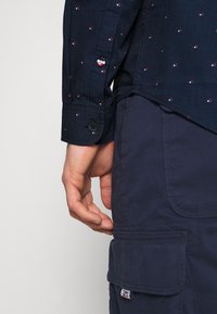 Gros plan d'une main près de la poche d'un pantalon cargo bleu marine et du poignet d'une chemise sombre à motifs à manches longues.
