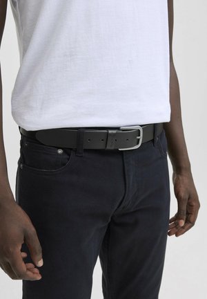 Zone de taille d'une personne portant une chemise blanche rentrée dans un pantalon noir avec une ceinture en cuir noire et une boucle en argent.