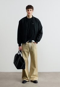 Νεαρός άνδρας που φοράει μαύρο μπουφάν και πουλόβερ με μπεζ παντελόνι φαρδιάς γραμμής, κρατώντας μαύρη τσάντα duffel, στέκεται μπροστά σε απλό φόντο.